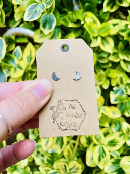 Hand holding tan tag with crescent moon earrings and "the beaded magpie" logo, against variegated green plant background.