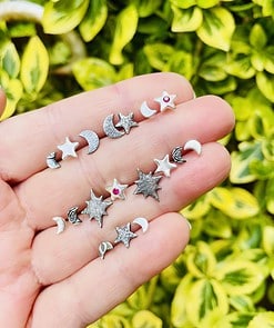 Hand holding small celestial-themed earrings with moons and stars, against a background of green and yellow foliage.