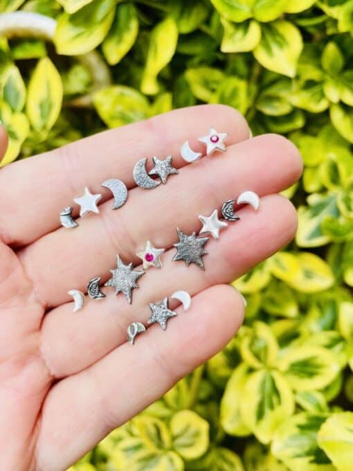 Hand holding small celestial-themed earrings with moons and stars, against a background of green and yellow foliage.