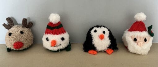 Four knitted plush toys: a reindeer, snowman, penguin, and Santa, lined up on a beige surface against a light blue wall.