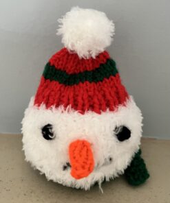 Knitted snowman ornament with red and green hat, black eyes, and an orange carrot nose on a beige surface.