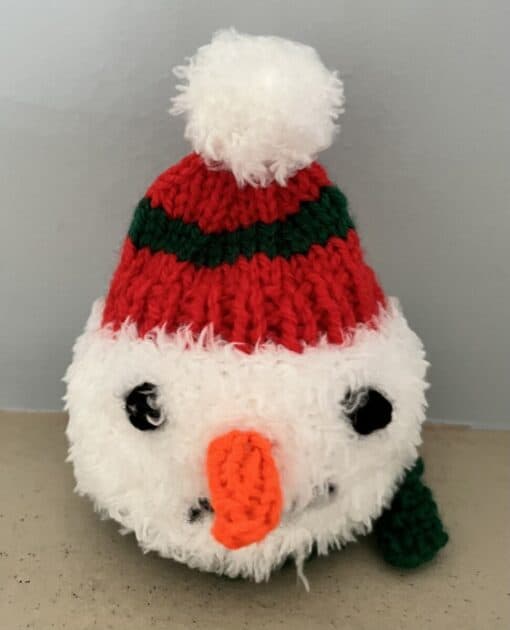 Knitted snowman ornament with red and green hat, black eyes, and an orange carrot nose on a beige surface.