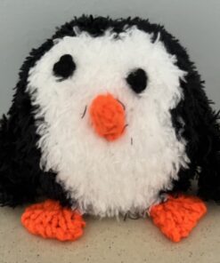 Fluffy knitted penguin toy with a white face, black body, orange beak and feet, sitting on a beige surface.