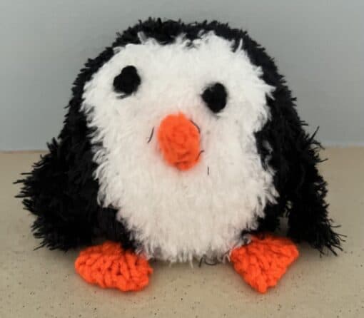 Fluffy knitted penguin toy with a white face, black body, orange beak and feet, sitting on a beige surface.