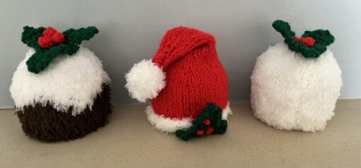 Knitted Christmas ornaments: two puddings and a Santa hat with holly decorations on a beige surface.