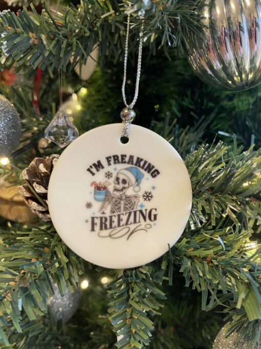 Ornament with a skeleton wearing a Santa hat and blue scarf, saying "I'm Freaking Freezing," hangs on a Christmas tree.