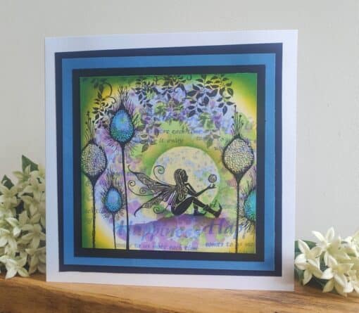 A fairy with wings sits on the ground in a colorful floral scene, framed with blue and black borders, accompanied by white flowers.