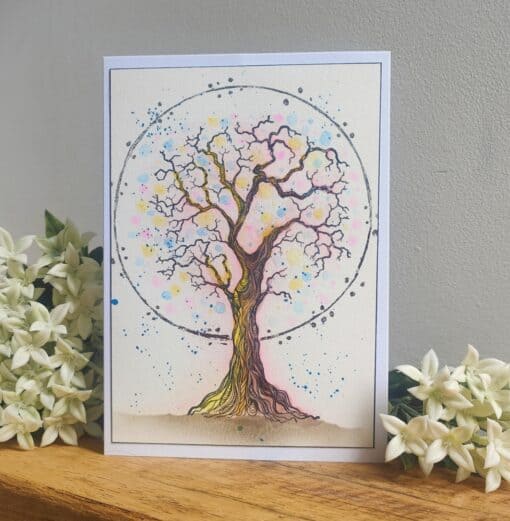 Watercolor painting of a tree with colorful leaves within a circle, surrounded by white flowers on a wooden surface.