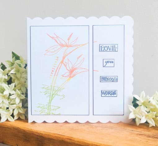 Greeting card on a wooden table featuring floral artwork and the words “Love you beyond words,” surrounded by white flowers.