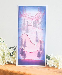 A greeting card with a pastel-colored landscape of mountains and pine trees, placed on a wooden surface with white flowers.