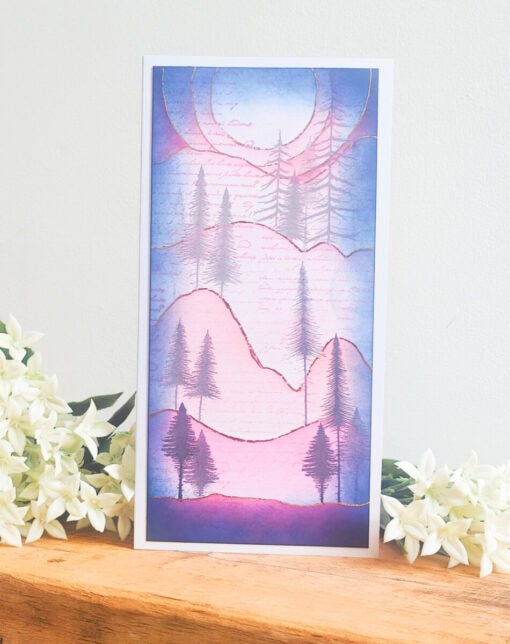 A greeting card with a pastel-colored landscape of mountains and pine trees, placed on a wooden surface with white flowers.