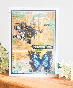 Handmade card featuring a bee, blue butterfly, and text "Live" and "Imagination." Card is on a wooden surface with white flowers.