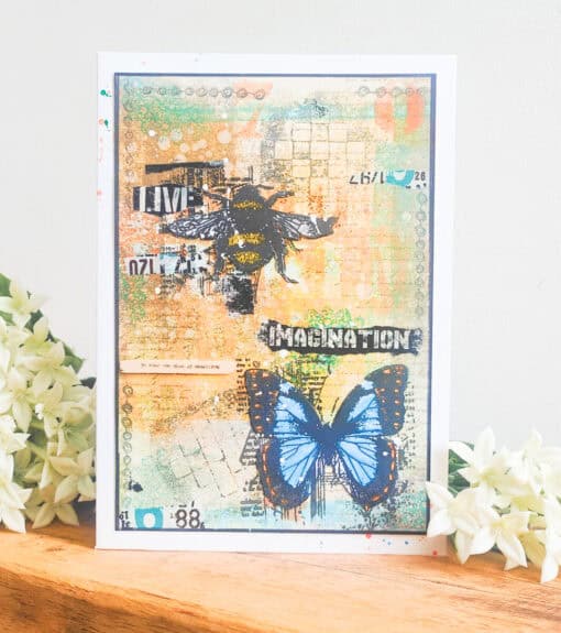 Handmade card featuring a bee, blue butterfly, and text "Live" and "Imagination." Card is on a wooden surface with white flowers.