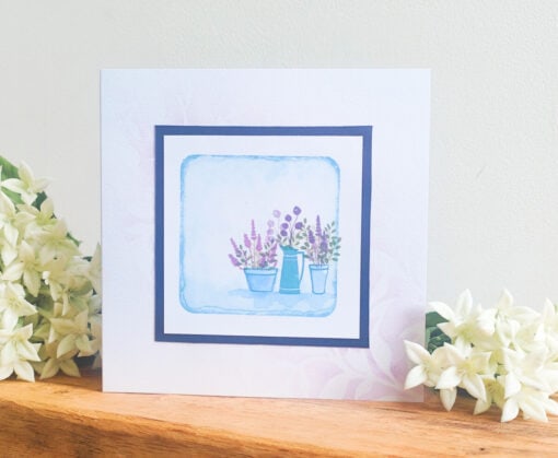 Greeting card with watercolor flowers in pots and a jug, bordered in black. It sits on a wooden surface with white flowers around.