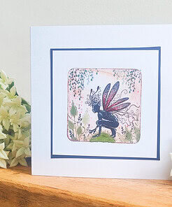 Greeting card featuring a fairy with red and blue wings, surrounded by flowers and foliage, placed on a wooden surface.