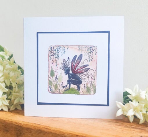 Greeting card featuring a fairy with red and blue wings, surrounded by flowers and foliage, placed on a wooden surface.