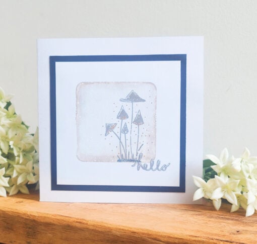 Greeting card with hand-drawn mushrooms and "hello" text, placed on a wooden surface with white flowers in the background.