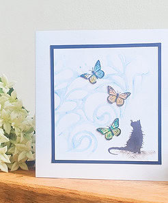 Handmade card with a black cat silhouette watching colorful butterflies, framed in blue with white flowers in the background.