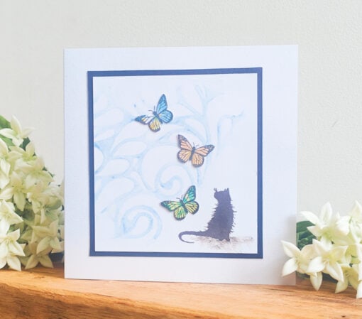 Handmade card with a black cat silhouette watching colorful butterflies, framed in blue with white flowers in the background.