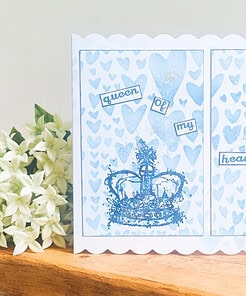 Greeting card with a blue crown, surrounded by hearts, and the text "queen of my heart." White flowers on a wooden surface.