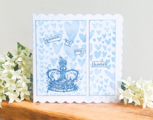 Greeting card with a blue crown, surrounded by hearts, and the text "queen of my heart." White flowers on a wooden surface.