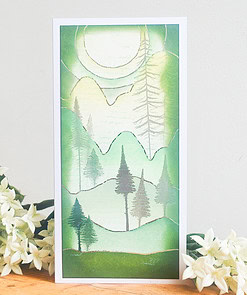 Handmade card featuring a serene green and white landscape with trees, mountains, and a large sun, placed beside white flowers.