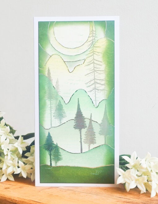 Handmade card featuring a serene green and white landscape with trees, mountains, and a large sun, placed beside white flowers.