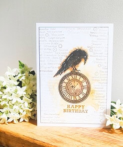 Birthday card featuring a black raven perched on a vintage clock, with "Happy Birthday" text, surrounded by white flowers.