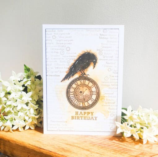 Birthday card featuring a black raven perched on a vintage clock, with "Happy Birthday" text, surrounded by white flowers.