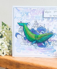 Handmade card with a green whale illustration, a "Whale you be mine?" message, and white flowers on a wooden shelf.