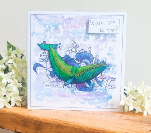 Handmade card with a green whale illustration, a "Whale you be mine?" message, and white flowers on a wooden shelf.