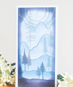 A greeting card with an artistic blue-toned mountain landscape and trees, placed on a wooden surface beside white flowers.