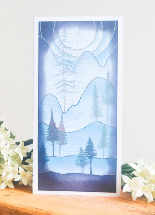 A greeting card with an artistic blue-toned mountain landscape and trees, placed on a wooden surface beside white flowers.