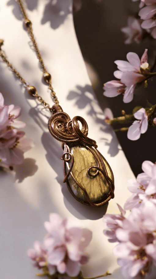 Wire-wrapped gemstone pendant necklace surrounded by pink flowers in soft focus.