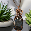 Green gemstone wrapped in intricate copper wire pattern, hanging on brown cord, with small potted succulent plants in background.