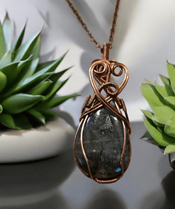 Green gemstone wrapped in intricate copper wire pattern, hanging on brown cord, with small potted succulent plants in background.