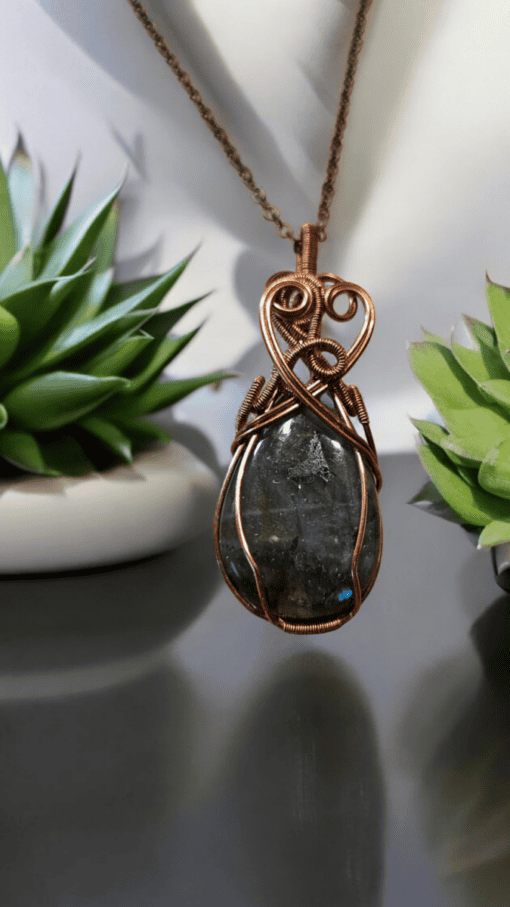 Green gemstone wrapped in intricate copper wire pattern, hanging on brown cord, with small potted succulent plants in background.