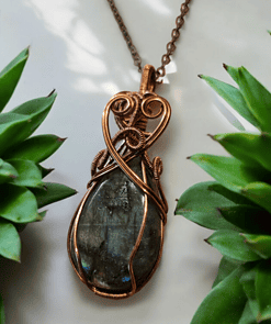 A copper-wrapped pendant with a dark stone center, hanging on a chain, surrounded by green succulent plants.