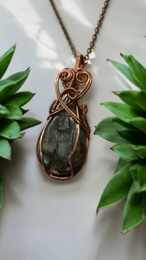 A copper-wrapped pendant with a dark stone center, hanging on a chain, surrounded by green succulent plants.