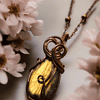 Gold and bronze pendant necklace with intricate wire wrapping, surrounded by soft-focused white flowers.