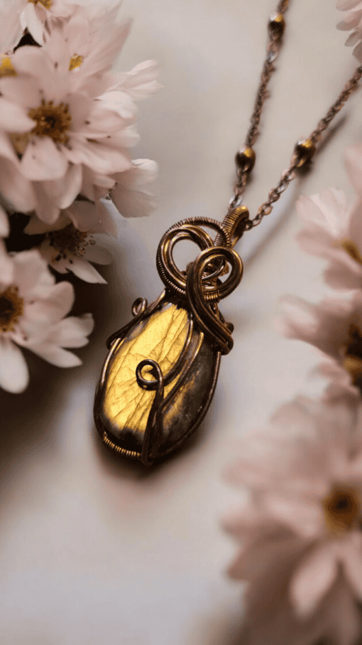 Gold and bronze pendant necklace with intricate wire wrapping, surrounded by soft-focused white flowers.