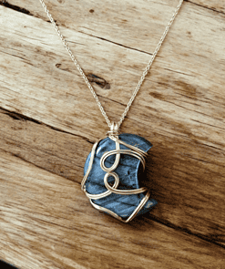 A blue stone pendant wrapped in gold wire on a thin gold chain, set against a rustic wooden surface.