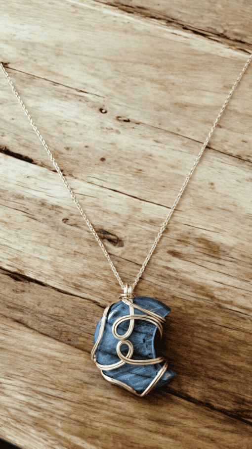 A blue stone pendant wrapped in gold wire on a thin gold chain, set against a rustic wooden surface.