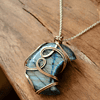 Blue labradorite stone wrapped in intricate gold wire, hanging from a silver chain, placed on a wooden surface.