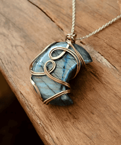 Blue labradorite stone wrapped in intricate gold wire, hanging from a silver chain, placed on a wooden surface.