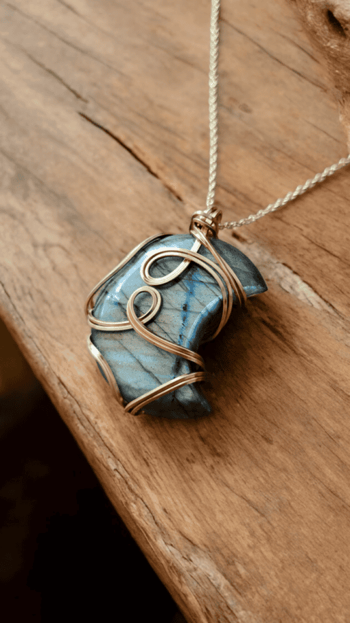 Blue labradorite stone wrapped in intricate gold wire, hanging from a silver chain, placed on a wooden surface.