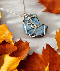 A blue gemstone pendant wrapped in silver wire rests among orange autumn leaves on a light gray surface.