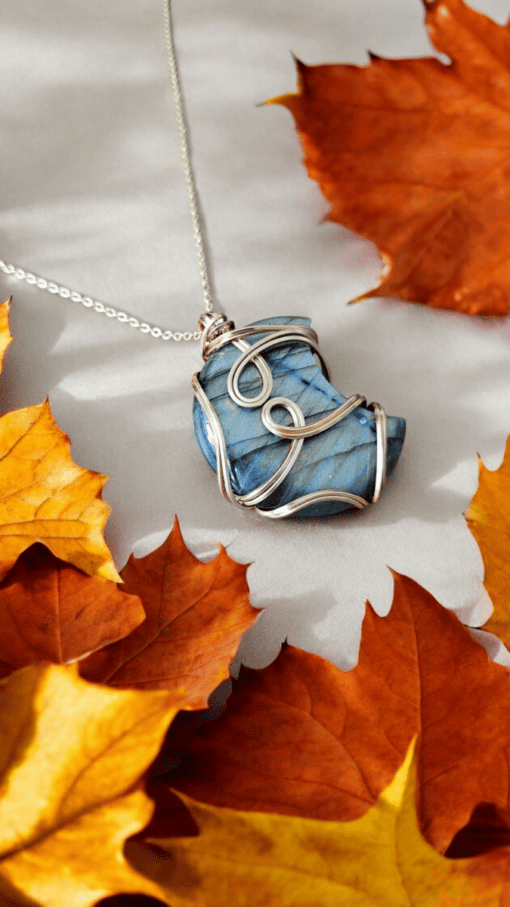 A blue gemstone pendant wrapped in silver wire rests among orange autumn leaves on a light gray surface.