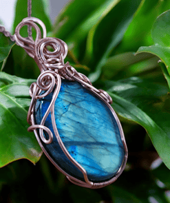 Boho-chic wire wrap pendant with a blue stone, featuring intricate wire wrapping, set against green leaves.