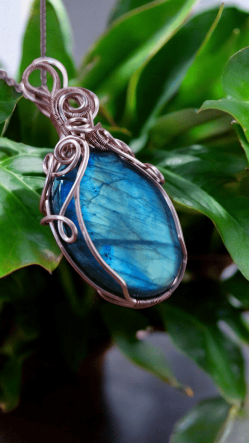 Boho-chic wire wrap pendant with a blue stone, featuring intricate wire wrapping, set against green leaves.